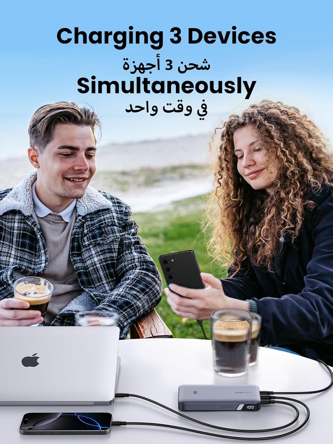 Two people sitting outdoors with a device charging three devices, including a laptop and smartphones.