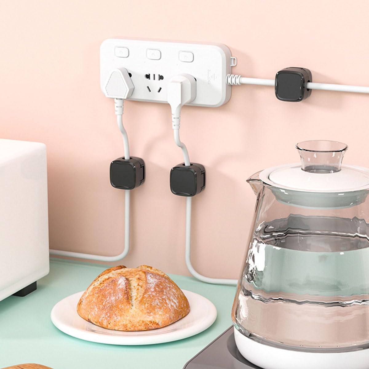 Wall-mounted power strip with multiple outlets and plugs, next to a loaf of bread and a glass teapot on a table.