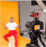 Camera on a tripod with a microphone and screen displaying a person in front of an orange backdrop.