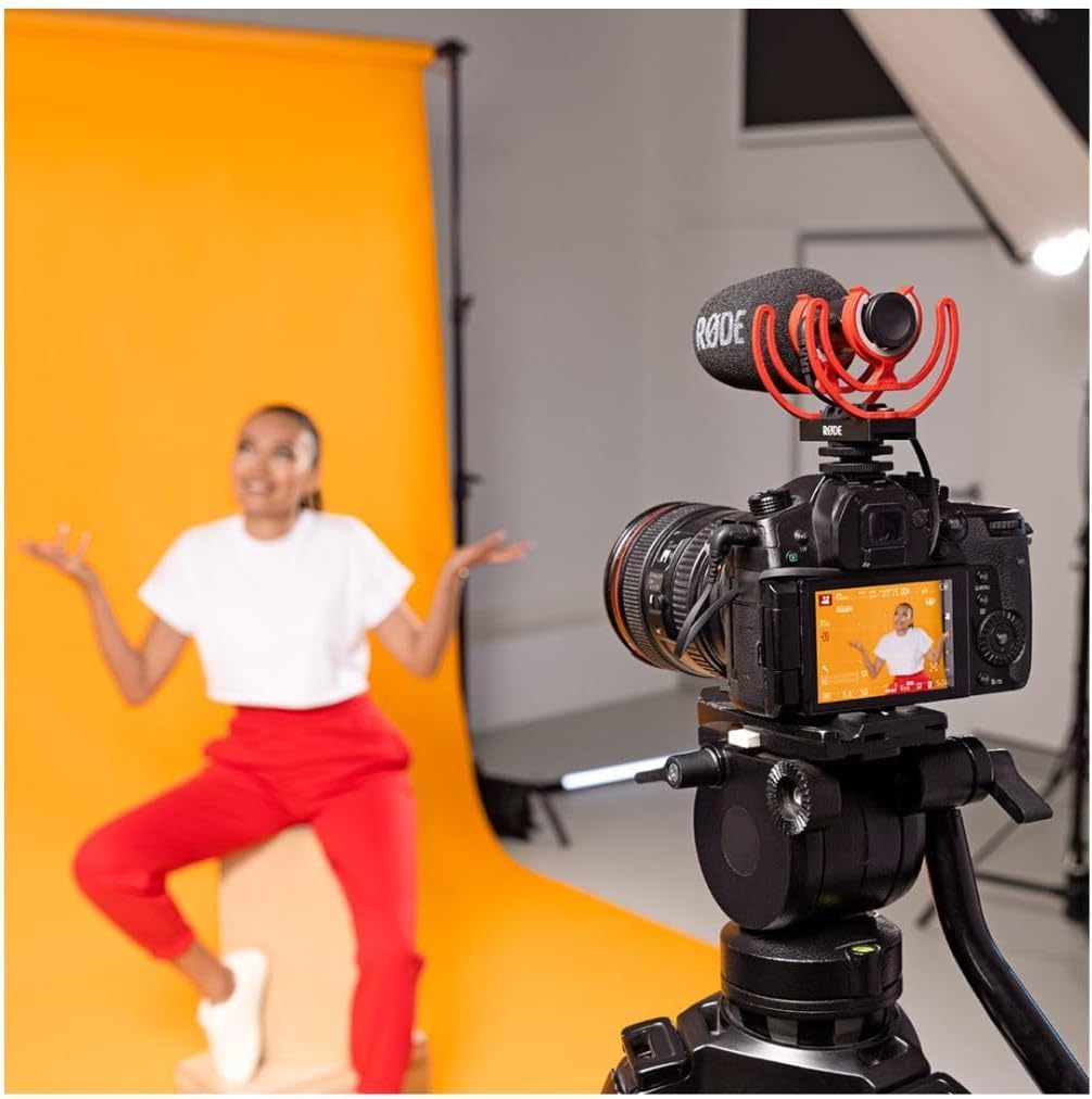Camera on a tripod with a microphone and screen displaying a person in front of an orange backdrop.