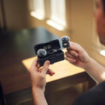 Person holding two small gray objects with a black case on a wooden surface.